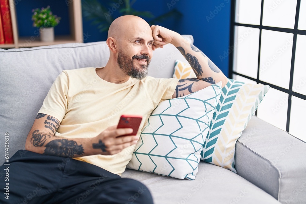 Young bald man using smartphone sitting on sofa at home Young bald man using smartphone sitting on sofa at home