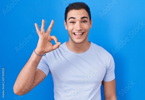 Young hispanic man standing over blue background smiling positive doing ok sign with hand and fingers. successful expression.