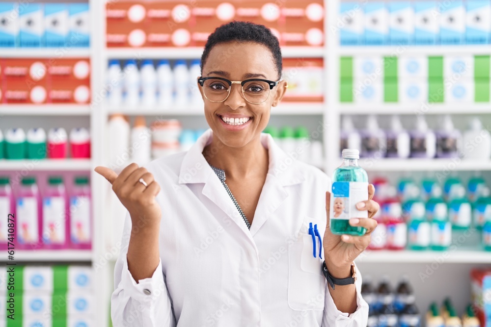 African american woman working at pharmacy drugstore holding syrup pointing thumb up to the side smiling happy with open mouth