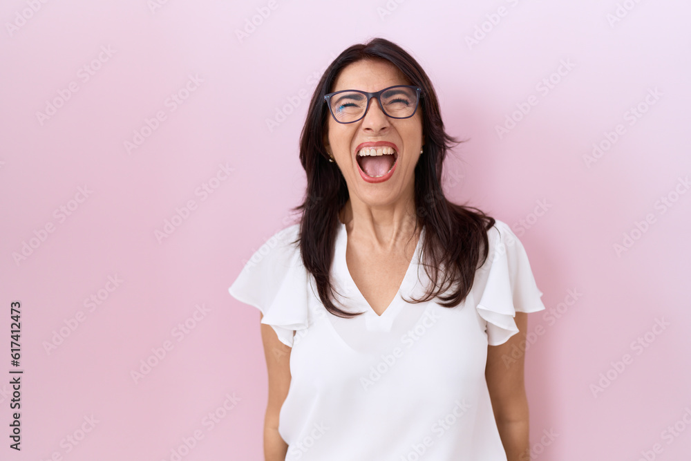 Middle age hispanic woman wearing casual white t shirt and glasses angry and mad screaming frustrated and furious, shouting with anger. rage and aggressive concept. Middle age hispanic woman wearing casual white t shirt and glasses angry and mad screaming frustrated and furious, shouting with anger. rage and aggressive concept.