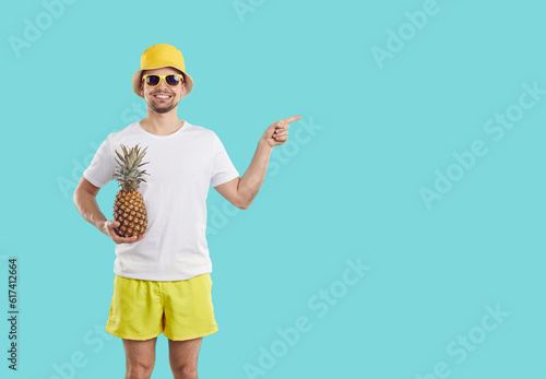 Cheerful man with pineapple on light blue background advertises new summer promotion or offers sale. Smiling man in shorts, T-shirt, panama and sunglasses points his finger at acopy space. Banner. photo