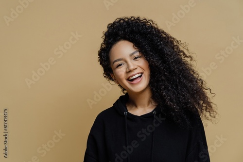 Positive African American woman smiles, tilts head happily, wearing black sweatshirt, isolated on beige background, radiating joy. Happiness personified.