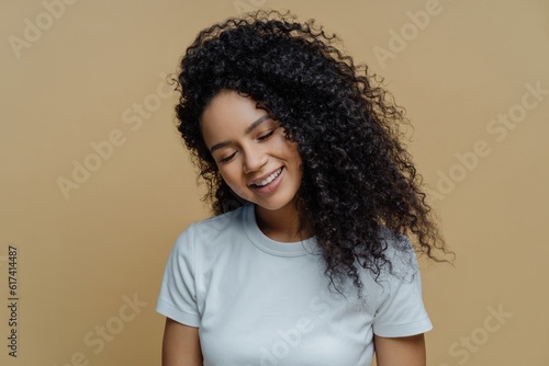 Pleased woman smiles, eyes closed, snow-white teeth, curly hair, casual t-shirt, beige background. Happiness, joy concept.