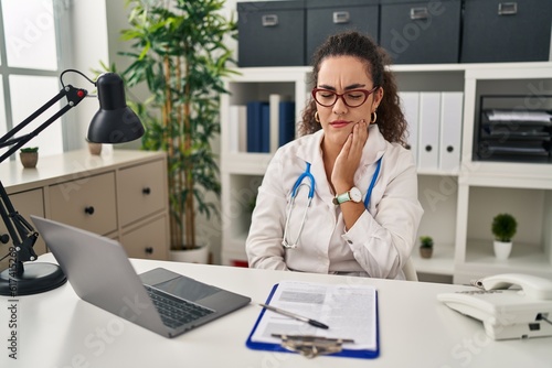 Young hispanic woman wearing doctor uniform and stethoscope touching mouth with hand with painful expression because of toothache or dental illness on teeth. dentist