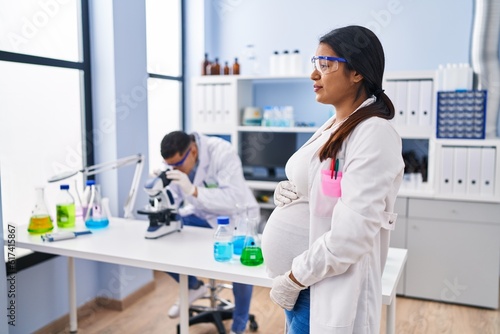 Young hispanic woman expecting a baby working at scientist laboratory looking to side, relax profile pose with natural face with confident smile.