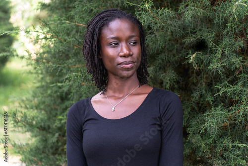 Young black woman with natural sister loc hairstyle smiling while standing in front of a pine tree at a park. No make up and wearing a black top