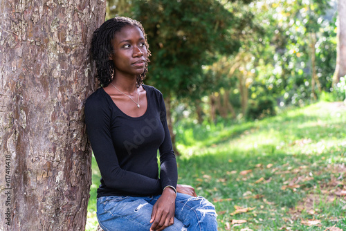 Young black woman with natural sister loc hair style sitting on a log in the park. Wearing blue jeans and a black top. No make up photo