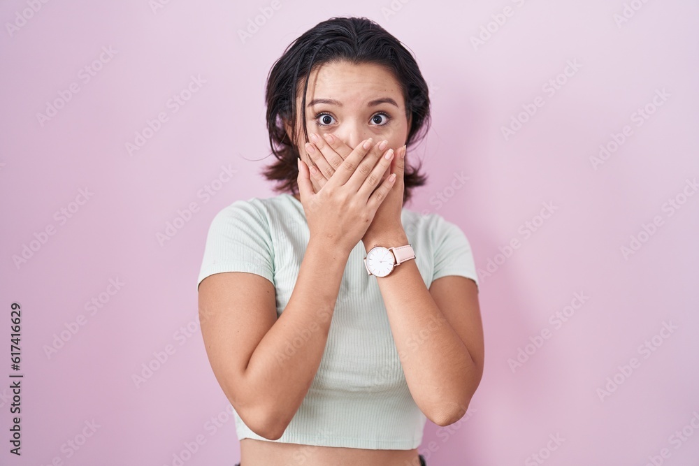 Hispanic young woman standing over pink background shocked covering mouth with hands for mistake. secret concept.