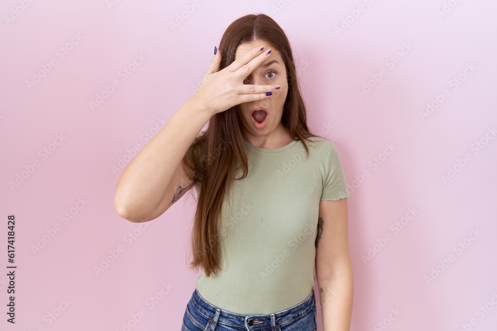 Beautiful brunette woman standing over pink background peeking in shock covering face and eyes with hand, looking through fingers with embarrassed expression. Beautiful brunette woman standing over pink background peeking in shock covering face and eyes with hand, looking through fingers with embarrassed expression.