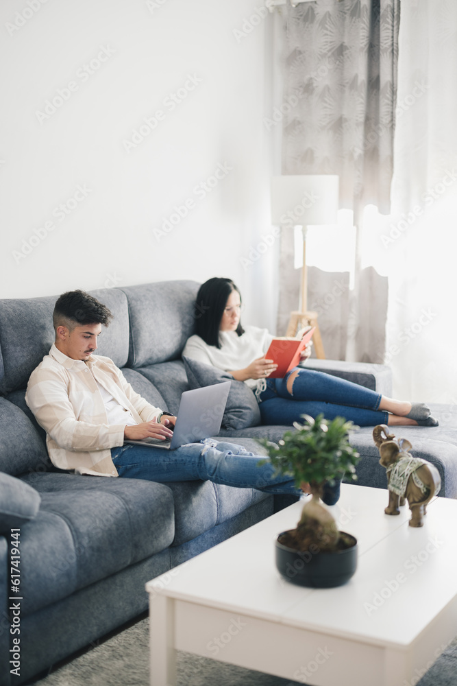 Young man working on sofa with laptop near woman reading a book Young man working on sofa with laptop near woman reading a book