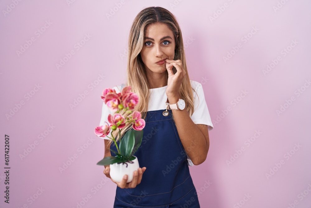 Young blonde woman wearing gardener apron holding plant mouth and lips shut as zip with fingers. secret and silent, taboo talking