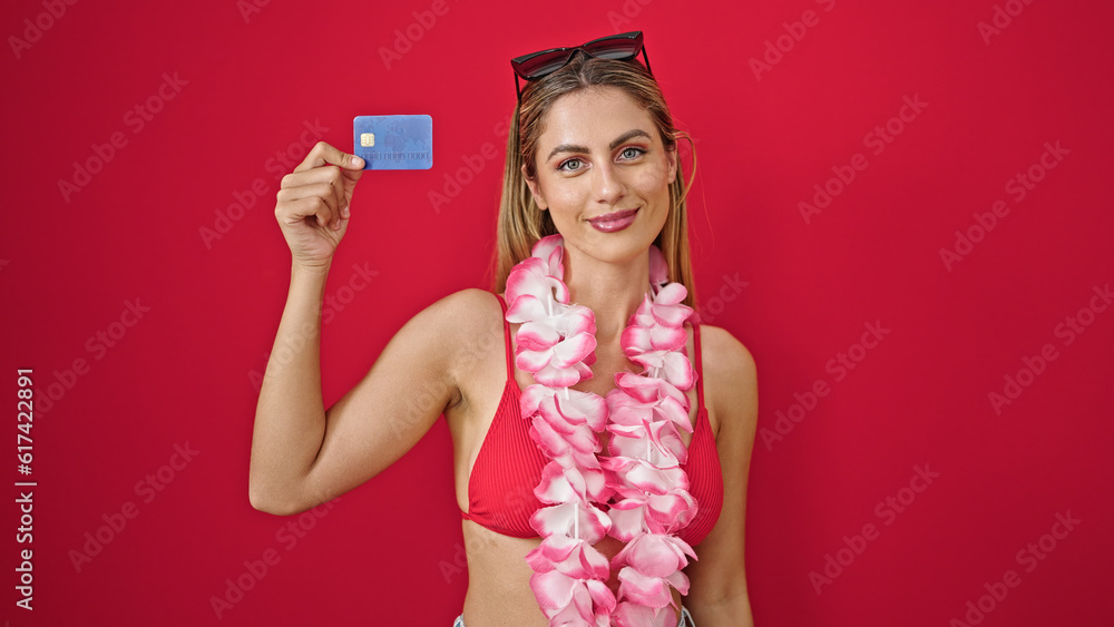Young blonde woman tourist holding credit card smiling over isolated red background Young blonde woman tourist holding credit card smiling over isolated red background