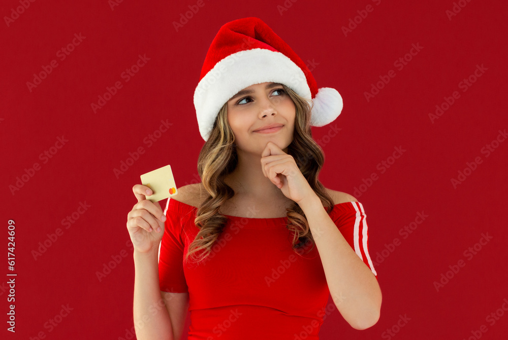 Merry teen girl wear casual red xmas dress Santa hat posing hold credit bank card look aside on area isolated on plain red background Happy New Year holiday concept Merry teen girl wear casual red xmas dress Santa hat posing hold credit bank card look aside on area isolated on plain red background Happy New Year holiday concept