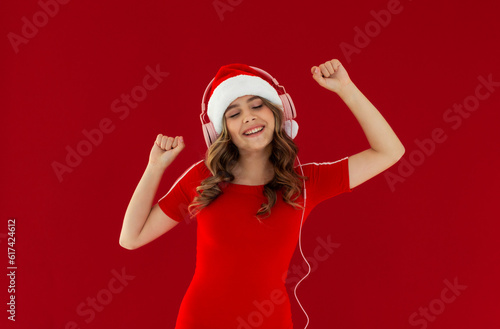 Funny young woman wearing green hoodie Christmas hat listening music with headphones keeping eyes closed isolated on red background studio portrait. Happy New Year celebration merry holiday concept