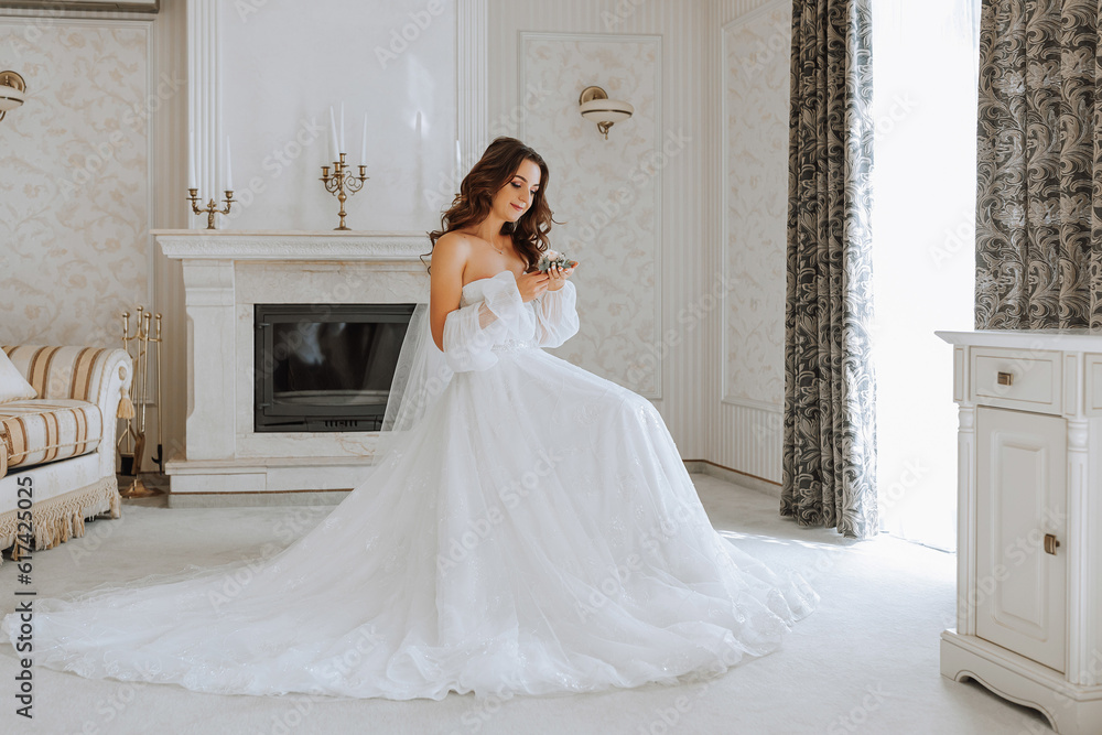 full-length portrait of the bride in the hotel room, the girl is sitting on a chair in a wedding dress in the middle of the room with a flower in her hands