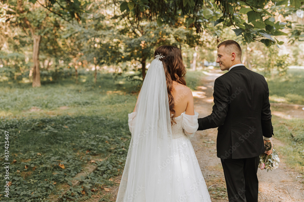 the bride and groom hold hands, walk in the garden looking at each other. Photo from the back the bride and groom hold hands, walk in the garden looking at each other. Photo from the back