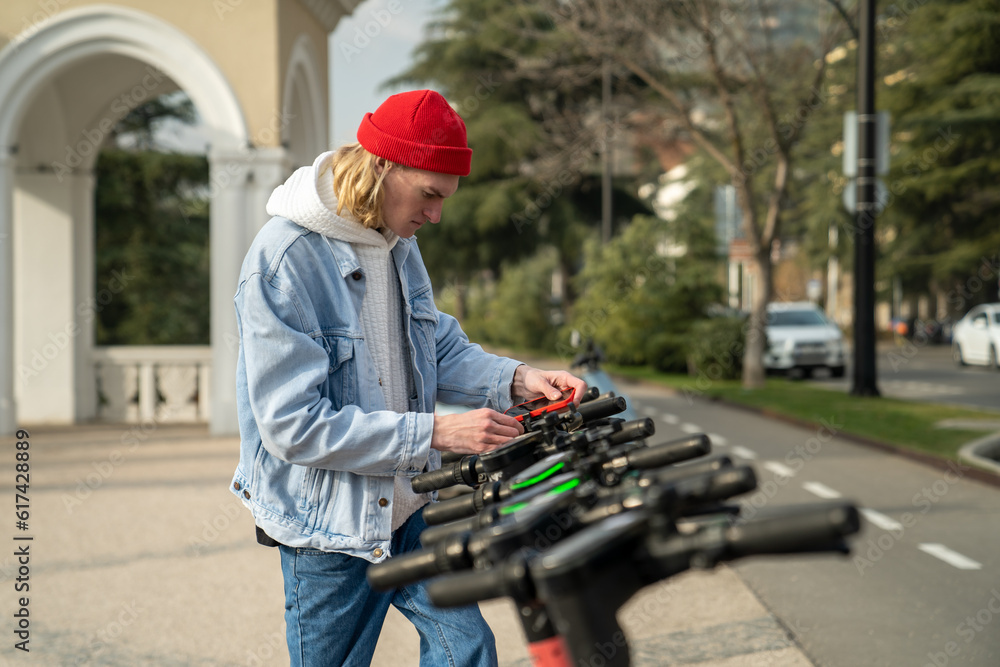 Young guy installs mobile phone on rented scooter.Man opens e-scooter using phone. Electric scooter is a new way of urban mobility. Green transport. Male rent eco transportation in summer Young guy installs mobile phone on rented scooter.Man opens e-scooter using phone. Electric scooter is a new way of urban mobility. Green transport. Male rent eco transportation in summer