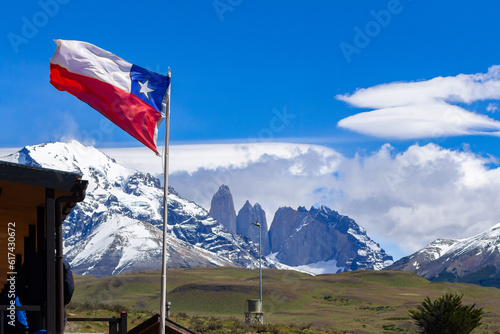 Paisagem da entrada do parque das Torres Del Paine com a casa principal no canto à esquerda, uma bandeira do chile tremulando ao vento e suas majestosas montanhas com neve e campos verdes. 