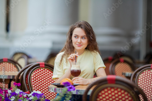 Beautiful dark-haired young curly woman sits in summer cafe and drinks coffee cocktail. Warm. Rest.