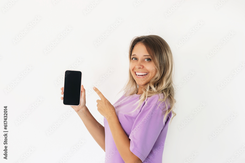 Young caucasian amazed smiling blonde woman in purple t-shirt holds mobile phone in her hand and points at black screen demonstrating copy space foror design on a white background. Mockup smartphone Young caucasian amazed smiling blonde woman in purple t-shirt holds mobile phone in her hand and points at black screen demonstrating copy space foror design on a white background. Mockup smartphone