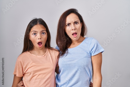 Young mother and daughter standing over white background in shock face, looking skeptical and sarcastic, surprised with open mouth