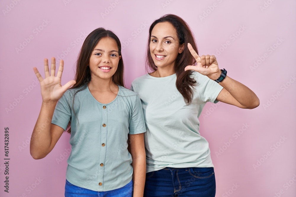 Young mother and daughter standing over pink background showing and pointing up with fingers number seven while smiling confident and happy. Young mother and daughter standing over pink background showing and pointing up with fingers number seven while smiling confident and happy.