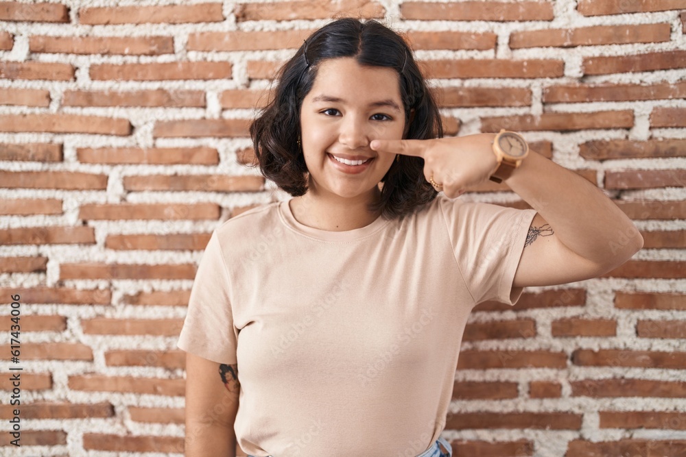 Young hispanic woman standing over bricks wall pointing with hand finger to face and nose, smiling cheerful. beauty concept Young hispanic woman standing over bricks wall pointing with hand finger to face and nose, smiling cheerful. beauty concept