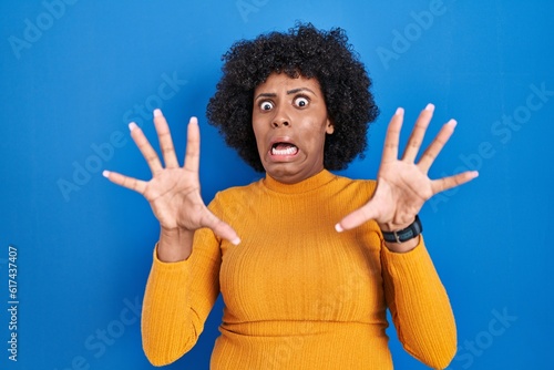 Φωτογραφία Black woman with curly hair standing over blue background afraid and terrified with fear expression stop gesture with hands, shouting in shock