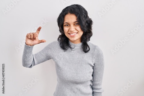 Hispanic woman with dark hair standing over isolated background smiling and confident gesturing with hand doing small size sign with fingers looking and the camera. measure concept.