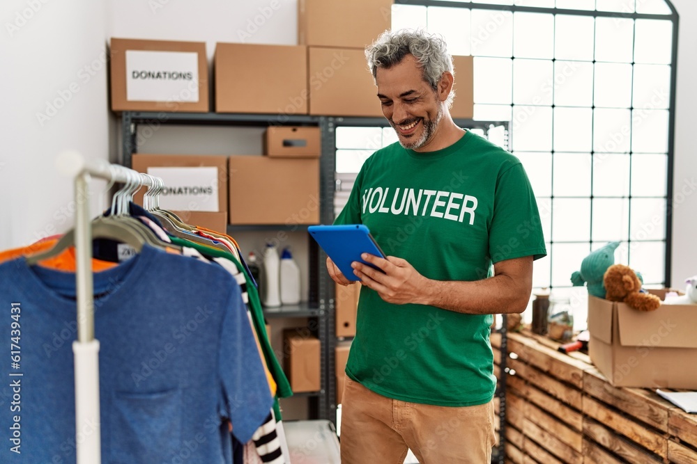 Middle age grey-haired man volunteer using touchpad holding clothes at charity center Middle age grey-haired man volunteer using touchpad holding clothes at charity center