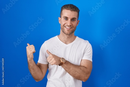 Young caucasian man standing over blue background pointing to the back behind with hand and thumbs up, smiling confident