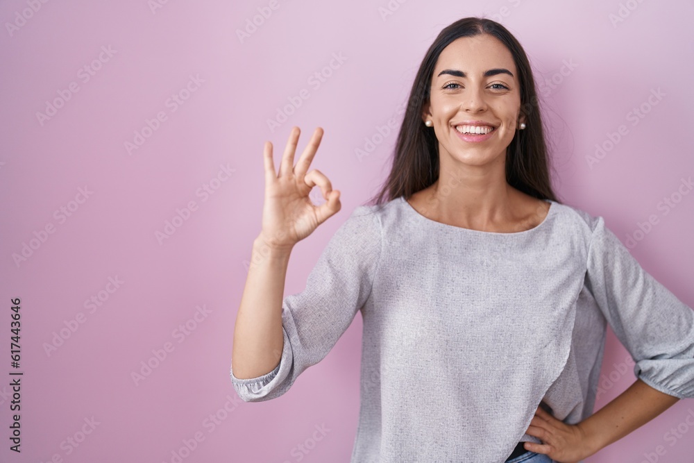 Young brunette woman standing over pink background smiling positive doing ok sign with hand and fingers. successful expression. Young brunette woman standing over pink background smiling positive doing ok sign with hand and fingers. successful expression.