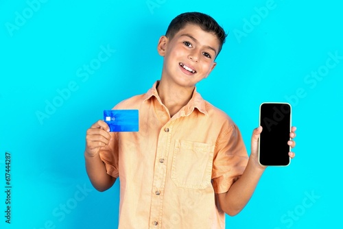 Little hispanic boy wearing orange shirt opened bank account, holding smartphone and credit card, smiling, recommend use online shopping application