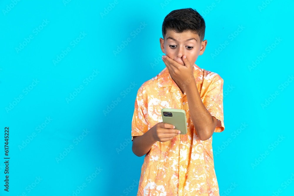 Little hispanic kid boy wearing hawaiian shirt being deeply surprised, stares at smartphone display, reads shocking news on website, Omg, its horrible! Little hispanic kid boy wearing hawaiian shirt being deeply surprised, stares at smartphone display, reads shocking news on website, Omg, its horrible!