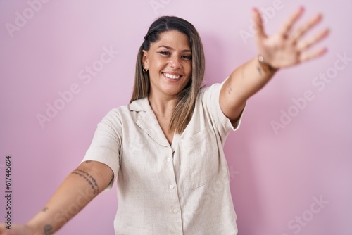Blonde woman standing over pink background looking at the camera smiling with open arms for hug. cheerful expression embracing happiness.