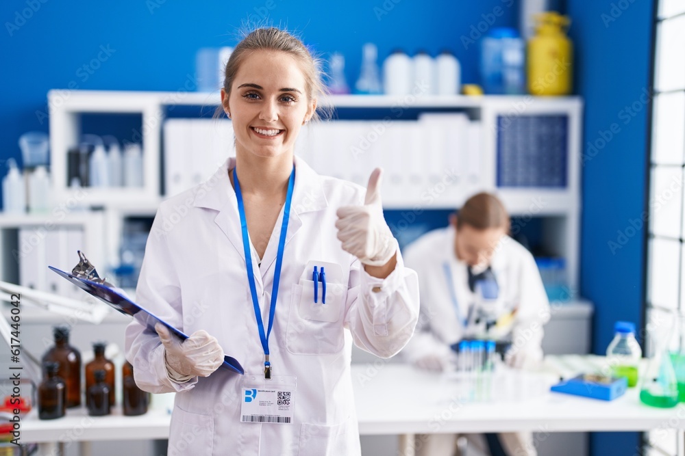 Blonde woman working at scientist laboratory smiling happy and positive, thumb up doing excellent and approval sign Blonde woman working at scientist laboratory smiling happy and positive, thumb up doing excellent and approval sign