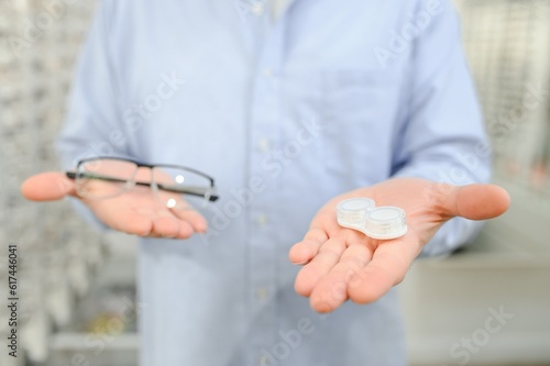 Ophthalmology Concept. Portrait of man choosing between eyeglasses and contact lenses standing in the optical store, holding plastic case and specs in both hands, looking at eyewear