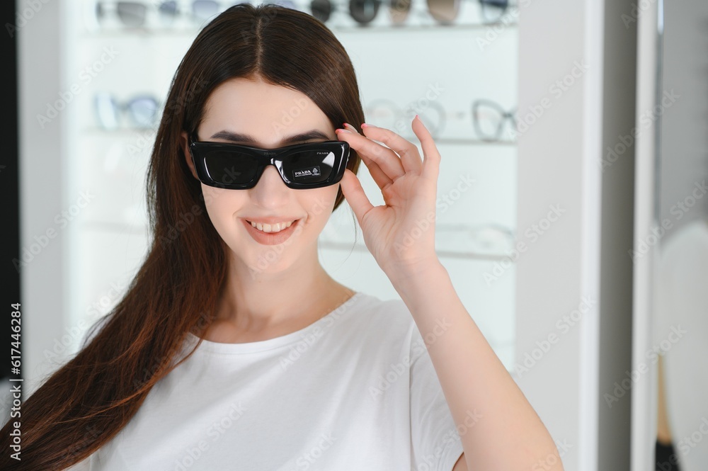 Portrait of a young woman shopping, standing in store and trying sunglasses near a mirror.