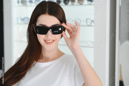 Portrait of a young woman shopping, standing in store and trying sunglasses near a mirror. © Serhii