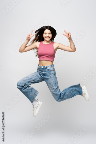 Smiling girl wearing pink top, gesturing, showing peace gesture, jumping isolated on gray background