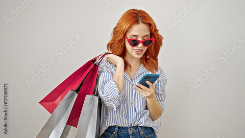 Young redhead woman using smartphone holding shopping bags over isolated white background
