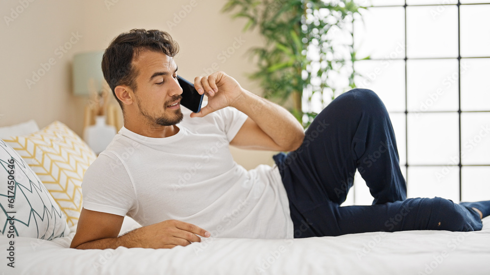 Young hispanic man talking on smartphone lying on bed at bedroom