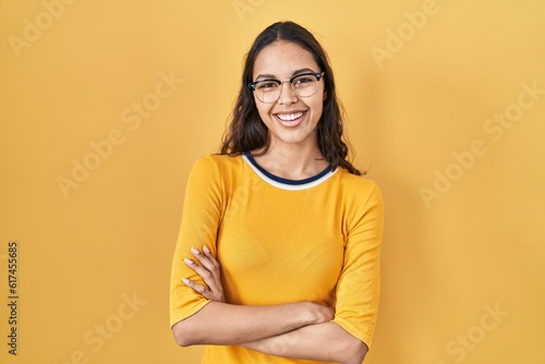 Young brazilian woman wearing glasses over yellow background happy face smiling with crossed arms looking at the camera. positive person.