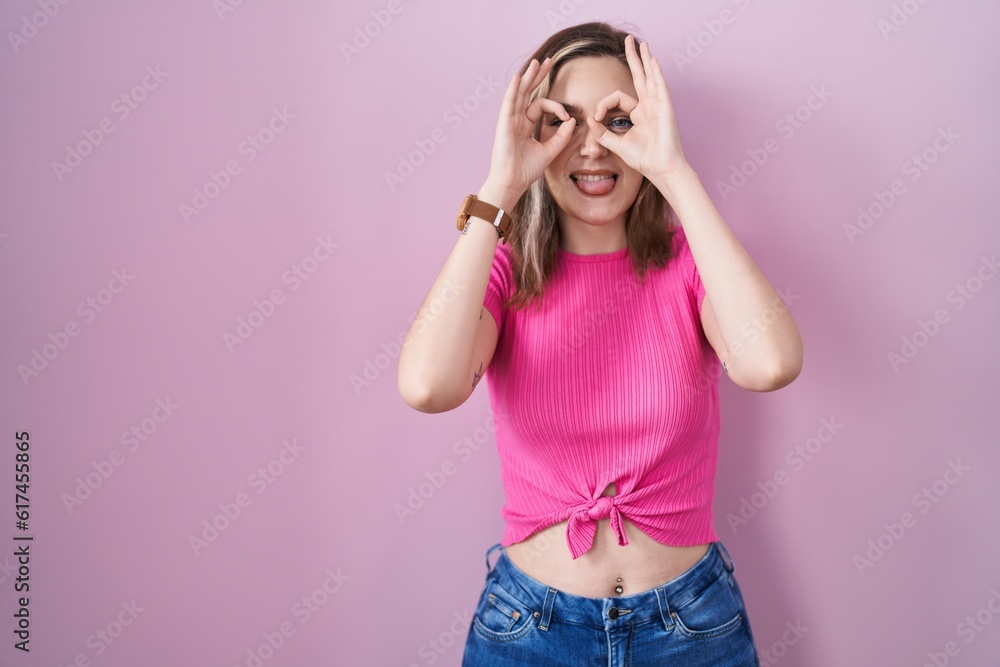 Blonde caucasian woman standing over pink background doing ok gesture like binoculars sticking tongue out, eyes looking through fingers. crazy expression. Blonde caucasian woman standing over pink background doing ok gesture like binoculars sticking tongue out, eyes looking through fingers. crazy expression.