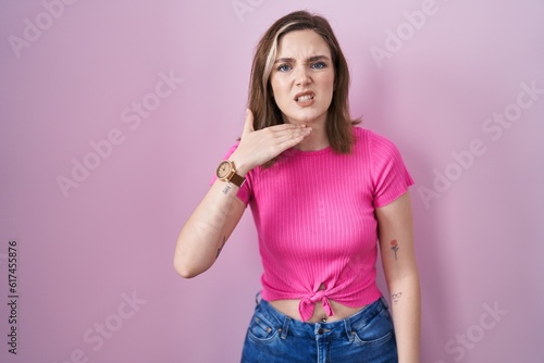 Blonde caucasian woman standing over pink background cutting throat with hand as knife, threaten aggression with furious violence