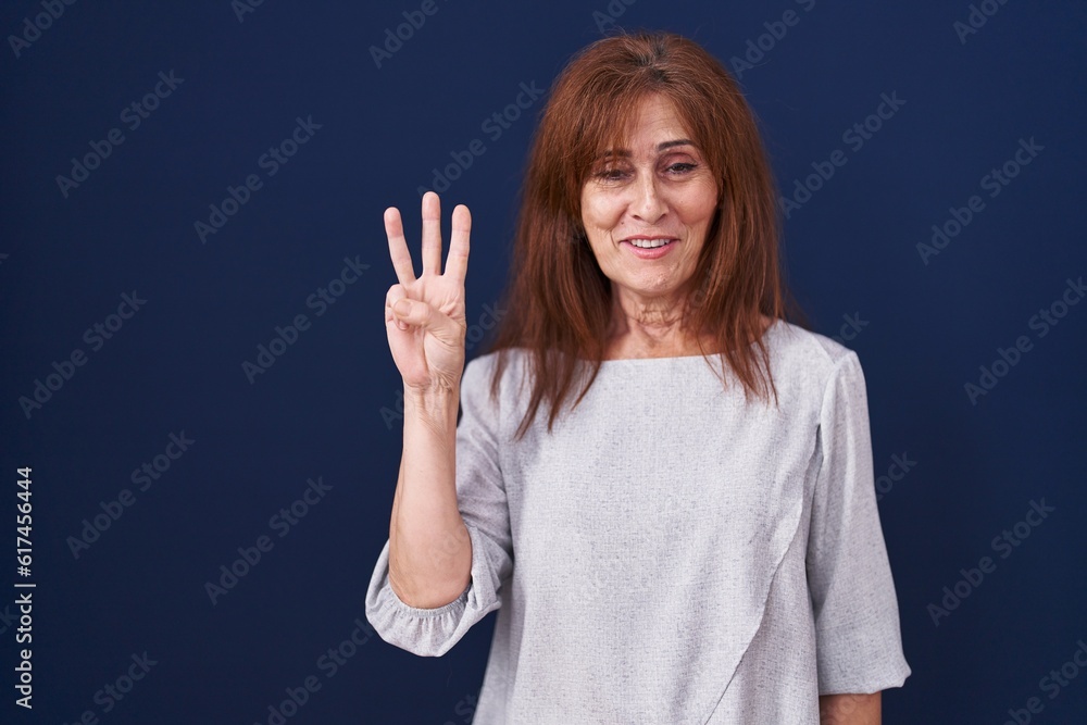 Middle age woman standing over blue background showing and pointing up with fingers number three while smiling confident and happy. Middle age woman standing over blue background showing and pointing up with fingers number three while smiling confident and happy.