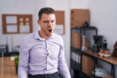 Young hispanic man at the office angry and mad screaming frustrated and furious, shouting with anger. rage and aggressive concept.