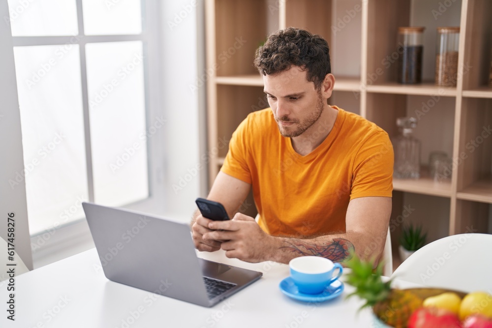 Young hispanic man using laptop and smartphone sitting on table at home Young hispanic man using laptop and smartphone sitting on table at home