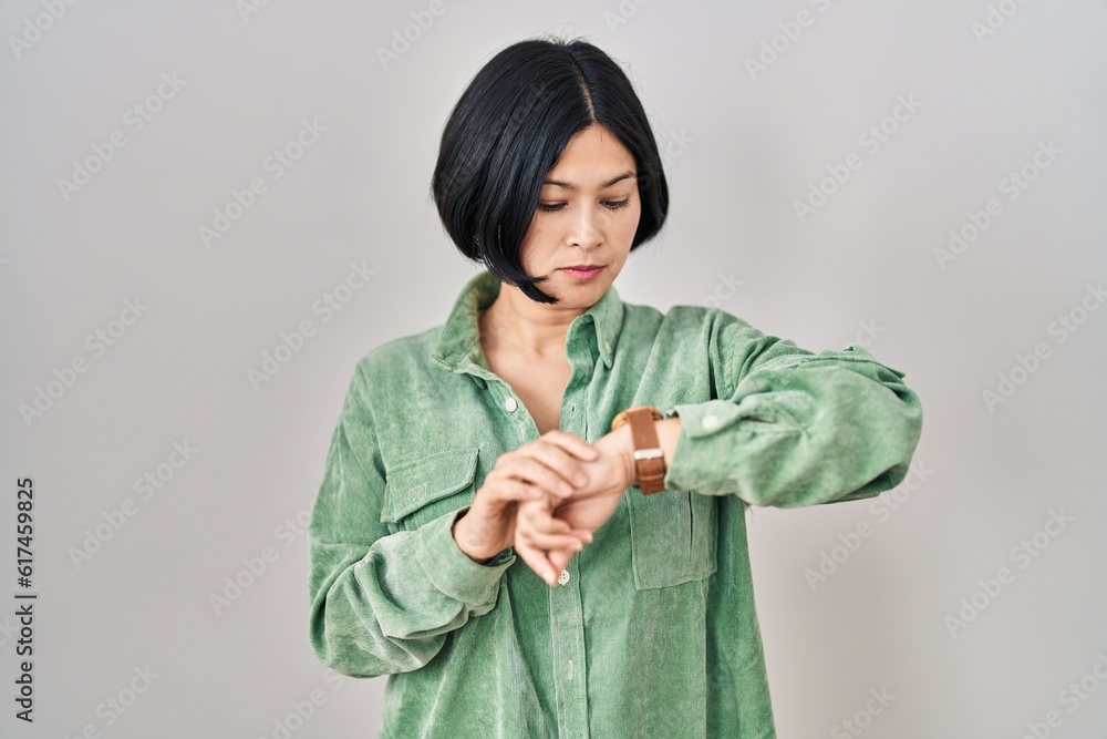Young asian woman standing over white background checking the time on wrist watch, relaxed and confident Young asian woman standing over white background checking the time on wrist watch, relaxed and confident