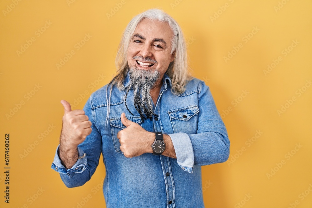Middle age man with grey hair standing over yellow background pointing to the back behind with hand and thumbs up, smiling confident Middle age man with grey hair standing over yellow background pointing to the back behind with hand and thumbs up, smiling confident